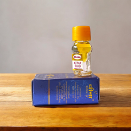 2ML glass bottle of OUD Attar by Veena, featuring a sleek gold cap and minimalist label, placed on a dark wooden surface with pieces of agarwood