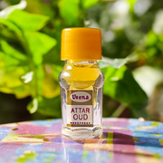 2ML glass bottle of OUD Attar by Veena, featuring a sleek gold cap and minimalist label, placed on a dark wooden surface with pieces of agarwood