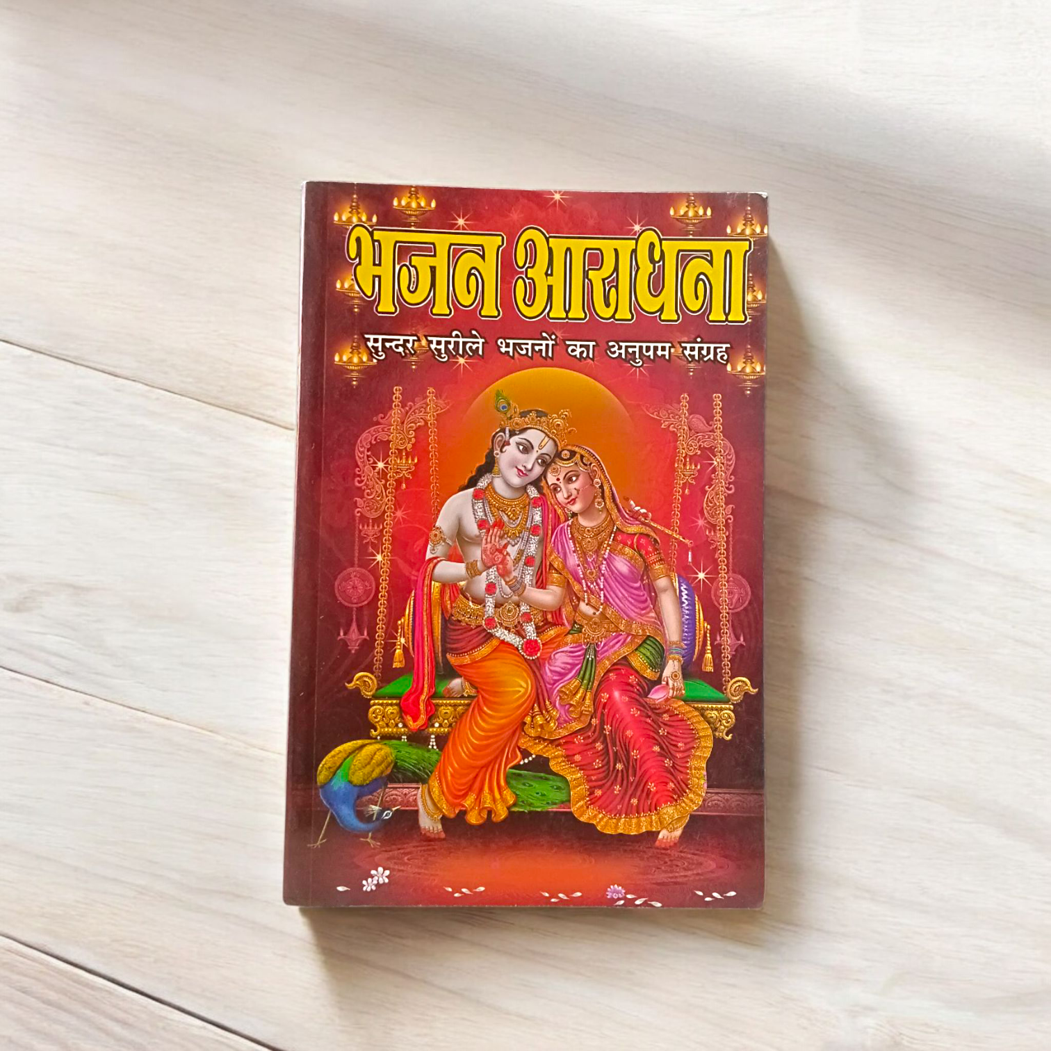 Bhajan Aradhana Hindi Book featuring colorful devotional cover, placed in a traditional worship setup with diya and flowers