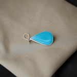 Close-up image of a natural Turquoise (Firoza) gemstone pendant with polished surface, paired with a black adjustable thread, displayed on a wooden background