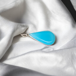 Close-up image of a natural Turquoise (Firoza) gemstone pendant with polished surface, paired with a black adjustable thread, displayed on a wooden background