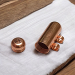 Five handcrafted cylindrical copper taweez placed beside Bhojpatra sheets on a clean, white background