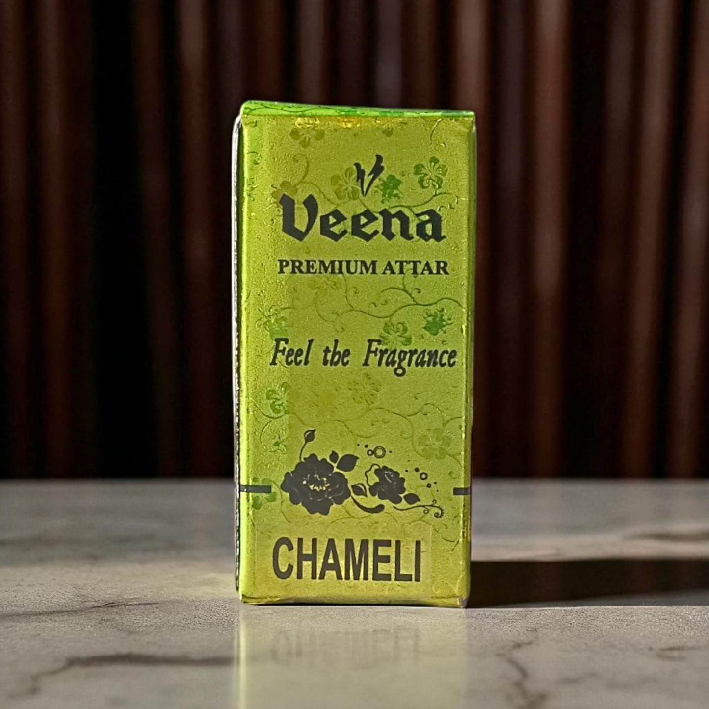 Glass bottle of 2ML Chameli Attar by Veena, adorned with a golden cap and floral-themed label, surrounded by jasmine flowers on a wooden surface